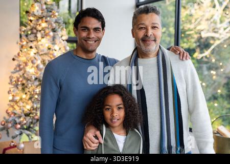 Three-generation family smiling before Christmas tree ornaments, lights and wrapped gifts at home Stock Photo