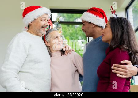 Diverse family standing in living room wearing Santa hats and antlers by Christmas tree and window Stock Photo