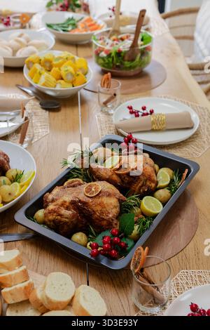Roast chickens are resting in black pan on wooden dining table, with citrus slices, rosemary sprigs Stock Photo