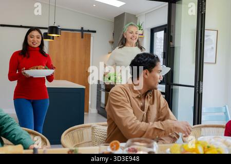 Diverse family members carrying roasted meat platter and salad bowl in kitchen, copy space Stock Photo