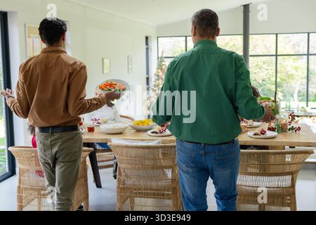 Diverse family carrying large salad bowls toward seated members at wooden dining table by windows Stock Photo