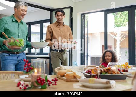 Diverse family carrying salad and roast chicken through folding doors into kitchen by festive table Stock Photo