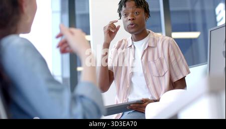 Discussing Black man holding tablet pointing at temple in office, with blurred coworker and monitor Stock Photo
