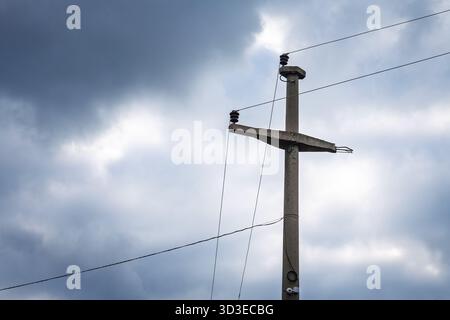 Close view of an old concrete utility pole with ceramic insulators and thin overhead lines against a cloudy sky. The worn fittings and simple single-p Stock Photo