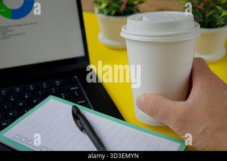 Morning workspace with takeaway coffee cup.Hand holding a takeaway coffee cup near laptop and notepad with pen on a bright desk. Stock Photo