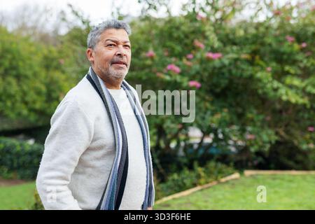 Mature adult man standing wearing light gray pullover sweater and long striped scarf in lush garden Stock Photo