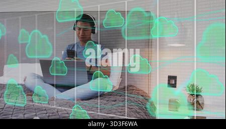 Sitting Asian man wearing headphones tapping smartphone and coding on laptop on bed, cloud overlay Stock Photo