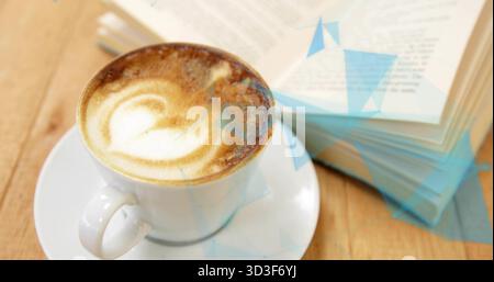 Sitting white ceramic cappuccino cup displaying heart foam art at cafe table, with paperback book Stock Photo