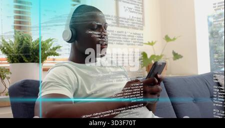 Scrolling phone, man in white T-shirt with over-ear headphones reading code overlay on blue sofa Stock Photo