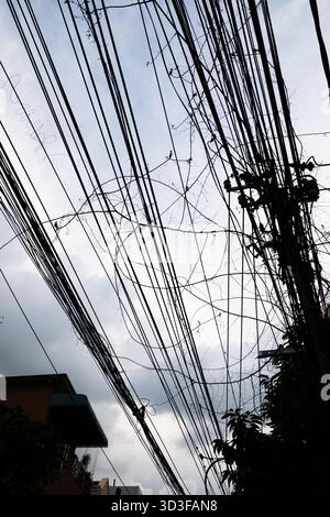 Silhouette of messy electrical wires, communication wires, and tree branch on blue sky in Bangkok, Thailand Stock Photo