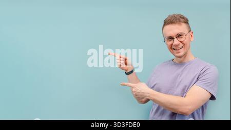 A cute funny young man in nerdy glasses points with his fingers at the blue background for the banner and text Stock Photo