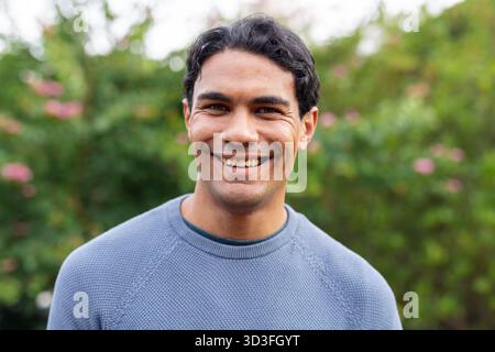 Man smiling while standing in garden in blue knit sweater among pink blossoms and foliage Stock Photo