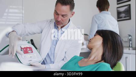 Dentist in white coat holding dental model and explaining to patient in clinic, with dental chair Stock Photo
