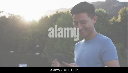 Smiling Asian man in light blue t-shirt holding smartphone on terrace, with coffee cup, copy space Stock Photo
