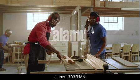 Two carpenters with safety goggles and ear protection cutting wooden board on table saw in workshop Stock Photo