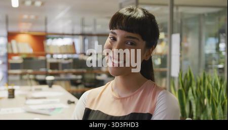Smiling woman with ponytail wearing colorblock top standing by desk in office with scattered papers Stock Photo