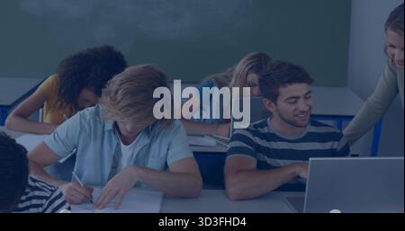 Instructor helping students at desks in classroom with laptops, notebooks and chalkboard background Stock Photo