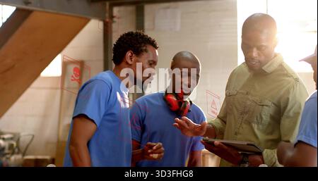 Examining tablet man in olive work shirt guiding blue tshirt workers in workshop with earmuffs Stock Photo