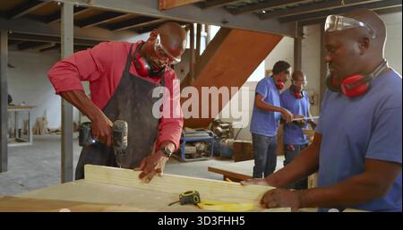 Drilling carpenter in red shirt securing plank at workshop bench, with cordless drill, copy space Stock Photo