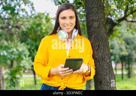 Smiling woman in yellow shirt holding tablet outdoors in park  Stock Photo