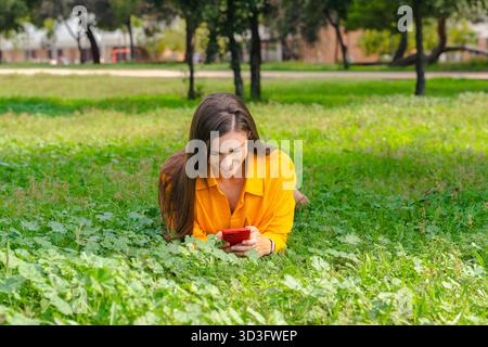 Smiling woman in yellow shirt lying on grass using red smartphone, green park. Stock Photo