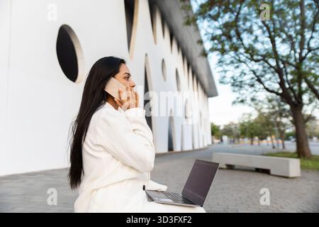 A businesswoman in a white modern urban setting is using a smartphone while working on her laptop. She represents modern connectivity and remote work Stock Photo