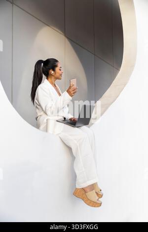 Businesswoman in a sleek white setting, multitasking with a laptop and smartphone. Modern urban environment highlights her connection and professional Stock Photo