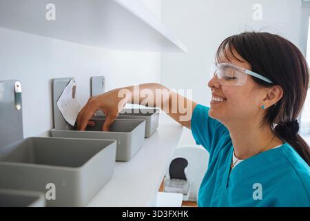 Side view of a cheerful female dental technician wearing protective glasses sorts through containers on a shelf in a dental laboratory, carefully orga Stock Photo