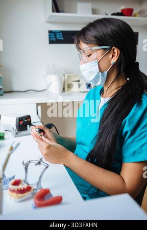 A female dental technician in a blue uniform is focused on crafting a dental prosthesis at her workstation. She wears safety goggles and a face mask w Stock Photo