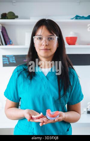 A female dental technician in blue scrubs and safety glasses holds dental prosthetics. She stands confidently in a dental laboratory, focused on her d Stock Photo