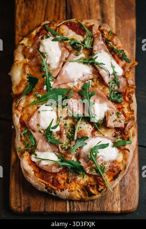 Savory Italian pizza topped with prosciutto, mozzarella, arugula, and basil pesto on a rustic wooden board. Perfect blend of flavors and textures for Stock Photo