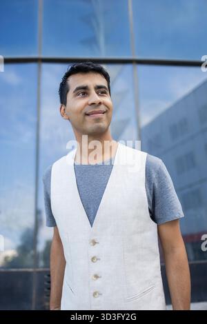 A young Hispanic man stands outdoors in the afternoon, gazing thoughtfully ahead. Dressed casually in a white vest and grey shirt, he appears relaxed Stock Photo