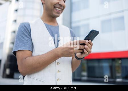Hispanic man smiling while using a smartphone outside a modern building. He is wearing a vest and a t-shirt, appearing relaxed and engaged with his mo Stock Photo