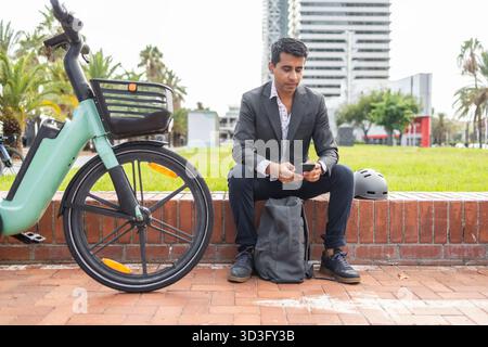 Hispanic man in formal attire sits on a brick ledge in an urban park. He is focused on his smartphone, a bicycle and helmet beside him, enjoying a mod Stock Photo