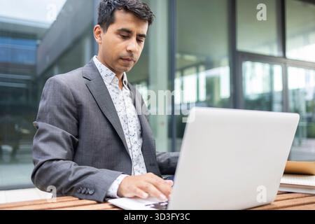Hispanic man in a suit sitting outside, focused on using his laptop in an outdoor setting during the afternoon. Represents modern business and digital Stock Photo