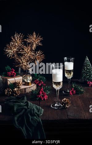 Two champagne flutes filled with bubbly are set on a wooden table, surrounded by festive decorations like pinecones, a snow globe, and gift boxes. A c Stock Photo