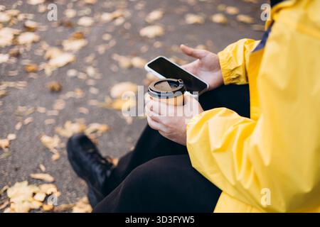 A man wearing a yellow raincoat sits outdoors in autumn, holding a smartphone in one hand and a cup of coffee in the other, surrounded by fallen leave Stock Photo