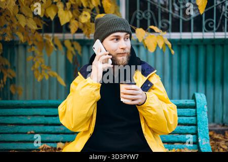 Man wearing a bright yellow raincoat sits on a bench surrounded by autumn leaves, holding a cup of coffee and talking on the phone, embracing the fall Stock Photo