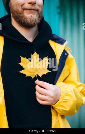A man dressed in a bright yellow raincoat holds a vibrant yellow maple leaf, capturing the essence of autumn. The scene is serene with the soft hue of Stock Photo