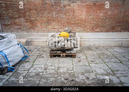 A yellow safety helmet rests on a pile of rough stones on a wooden pallet in front of a weathered brick wall, symbolizing ongoing construction work. Stock Photo