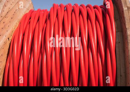Close-up of red electrical cables wound around a large wooden spool against a clear blue sky, essential for modern construction and infrastructure pro Stock Photo