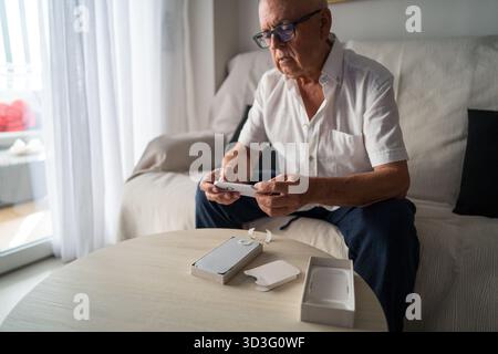 An elderly man sits on a comfortable sofa, eagerly unboxing a new smartphone designed for seniors. He examines the device with curiosity and interest, Stock Photo