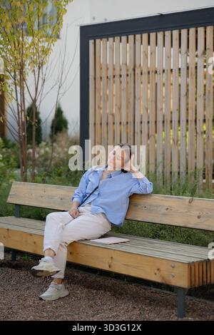 A young woman in a blue shirt and white pants relaxes on a wooden bench in a modern city park. The scene conveys tranquility and the beauty of urban o Stock Photo