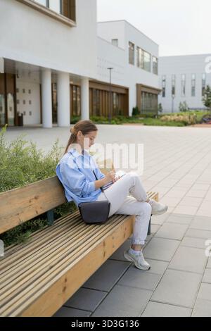 A young woman sits on a wooden bench in a modern city environment, capturing thoughts in a notebook. The setting is spacious, with contemporary archit Stock Photo