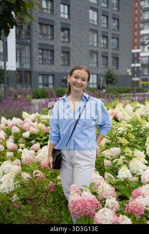 A young woman stands smiling amid vibrant flowers in a modern city setting, exuding confidence and joy. Urban buildings provide a dynamic backdrop for Stock Photo