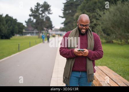 A smiling Latin American Cuban man enjoys a peaceful walk in a southern Chile park, using his smartphone to browse, surrounded by nature's serenity. Stock Photo