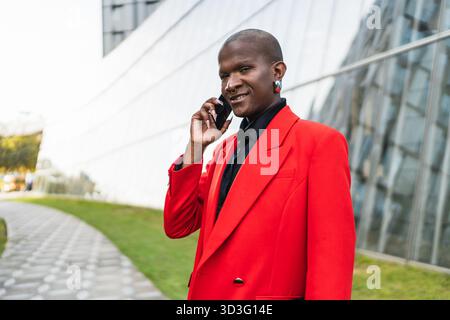 Young LGBTQ man in a vibrant red blazer holding a phone and smiling, standing confidently outside an office building, representing modern business and Stock Photo