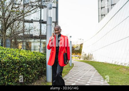 A young LGBTQ man wearing a stylish red blazer walks confidently outdoors, balancing a black folder while engaging in a conversation on his smartphone Stock Photo