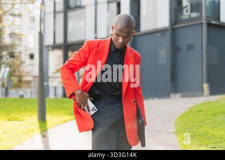 A fashionable young LGBTQ man in an orange blazer walking outdoors, confidently holding a smartphone. The modern background adds a professional touch. Stock Photo