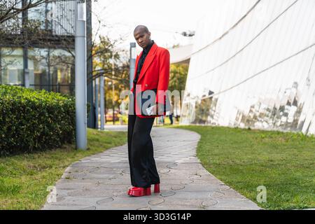 A young LGBTQ man confidently stands outside showcasing bold fashion. His bright red jacket and matching shoes highlight his unique style against a mo Stock Photo